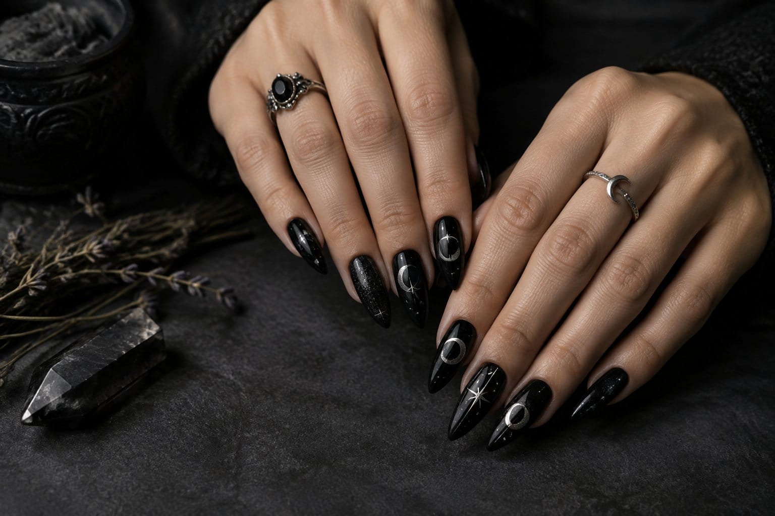 A pair of hands with black manicured nails rests beside a crystal and dried herbs.